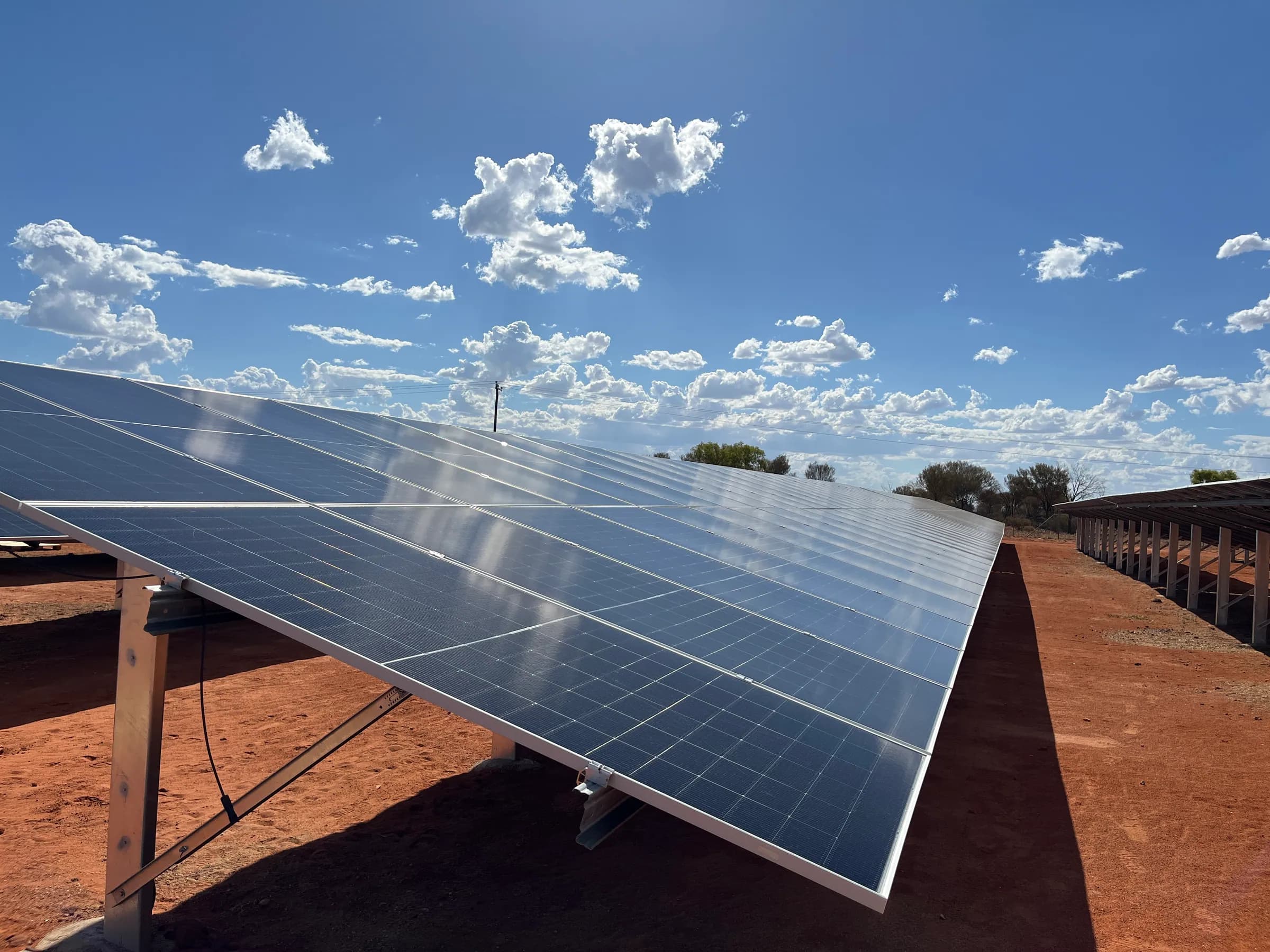 Long rows of ground-mounted solar panels on red earth under a blue sky with scattered cloud.
