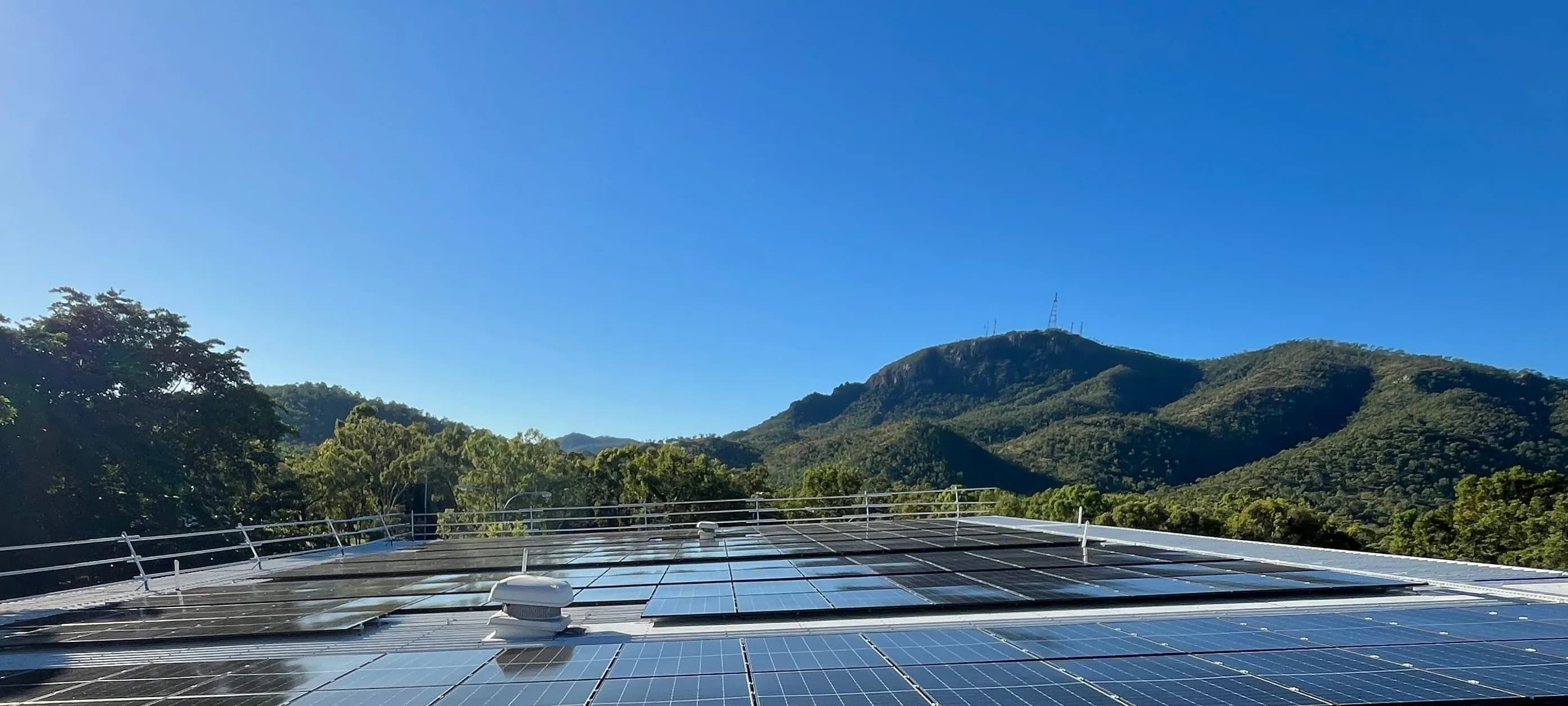 A rooftop solar array with forested hills behind it under a clear blue sky.